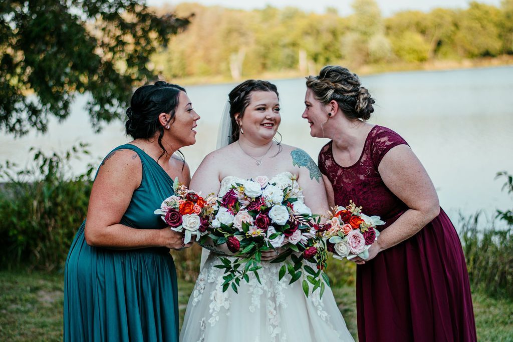 Three people in dresses smiling outdoors, holding a large bouquet by the water.