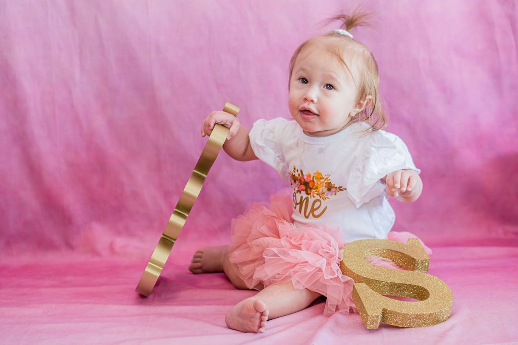 Baby in pink outfit sitting on a pink backdrop beside gold number decorations