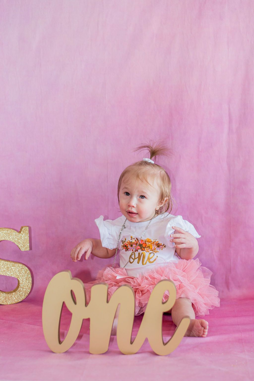 Baby seated against pink backdrop beside gold “one” sign, wearing a pink tutu and white shirt.