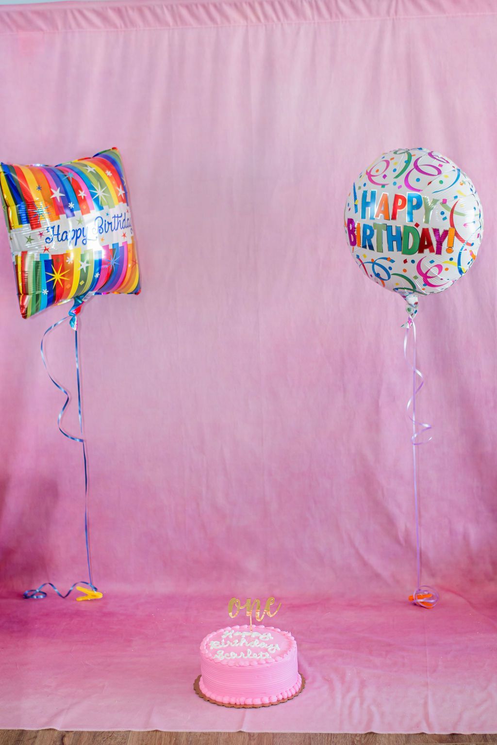Pink birthday backdrop with balloons and a small pink cake on the floor.