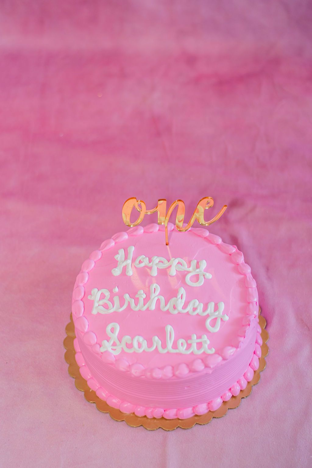 Pink birthday cake with gold “one” topper and “Happy Birthday Scarlett” icing on a pink background