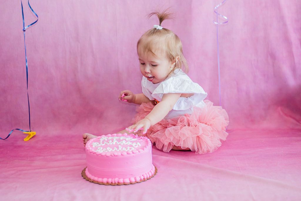 Toddler in a pink tutu reaching toward a pink frosted cake against a pink backdrop