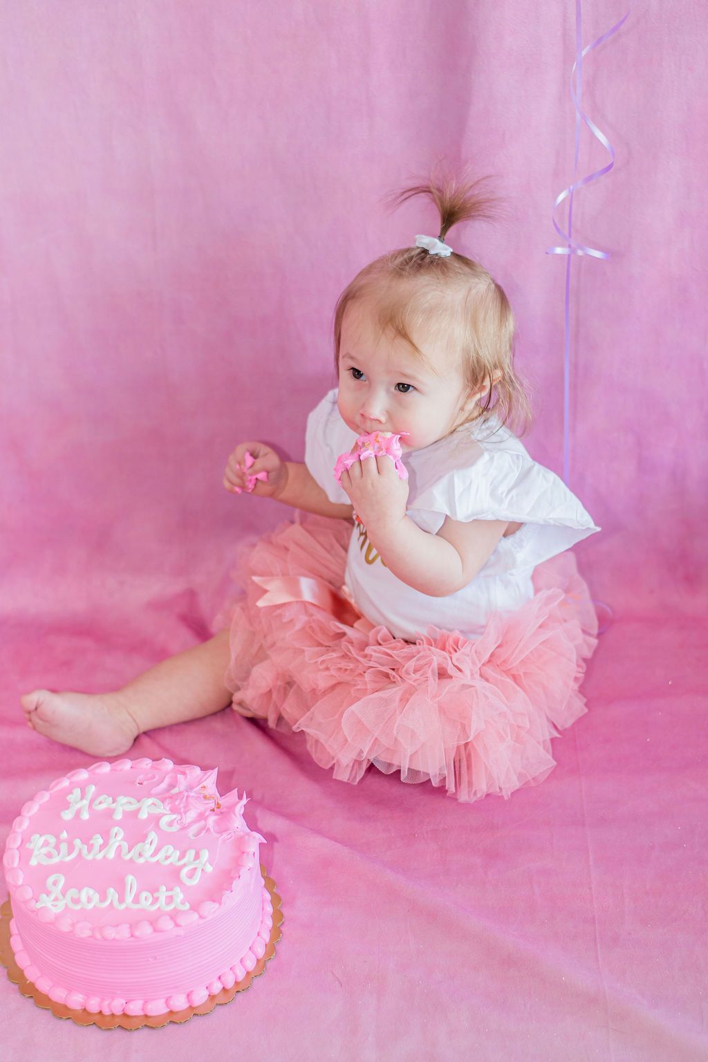 Toddler in a pink dress sitting beside a pink birthday cake with candles on a pink backdrop