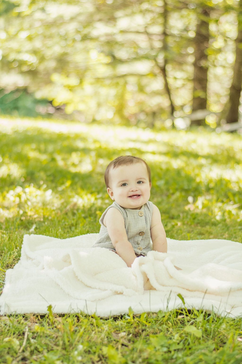 Baby sitting on a white blanket in a sunlit grassy park, smiling at the camera.
