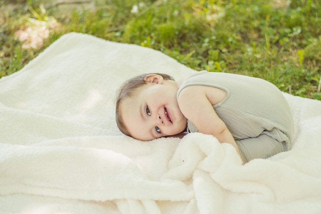 Baby lying on a white blanket in soft outdoor grass, smiling gently.