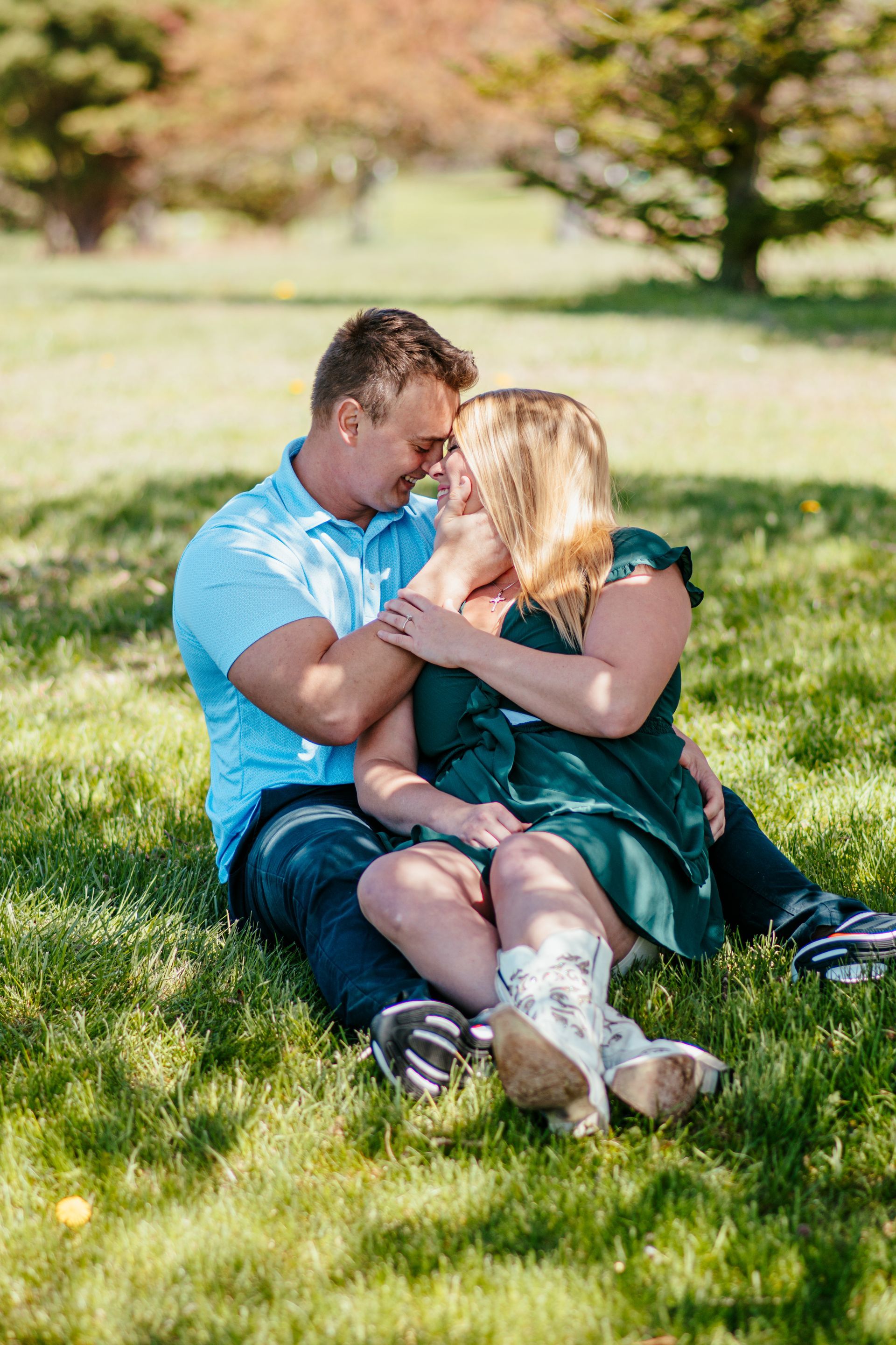 Couple sitting on grass in a park, embracing and smiling in sunlight.