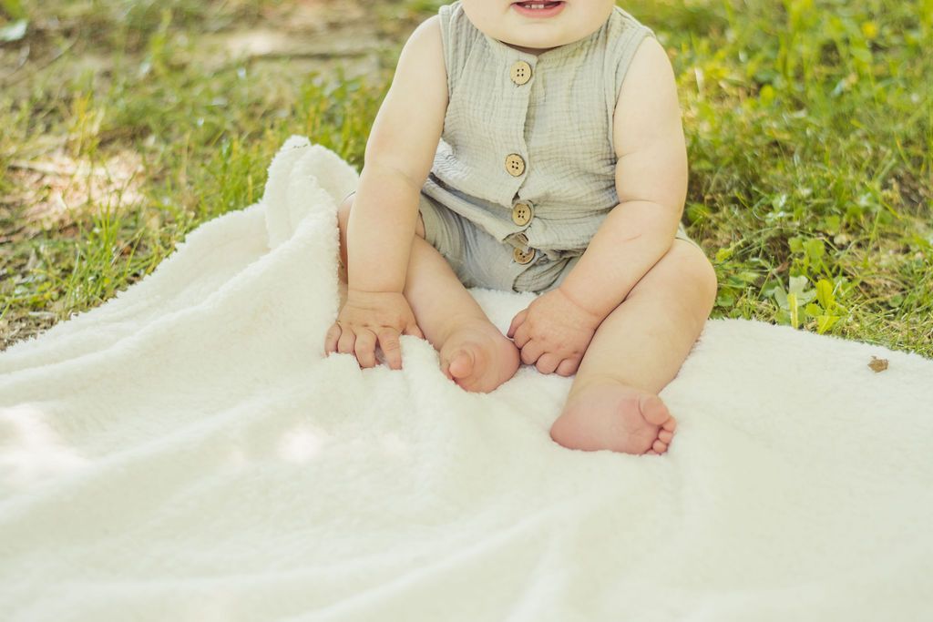 Baby sitting on a white blanket outdoors on grass, wearing a light gray outfit