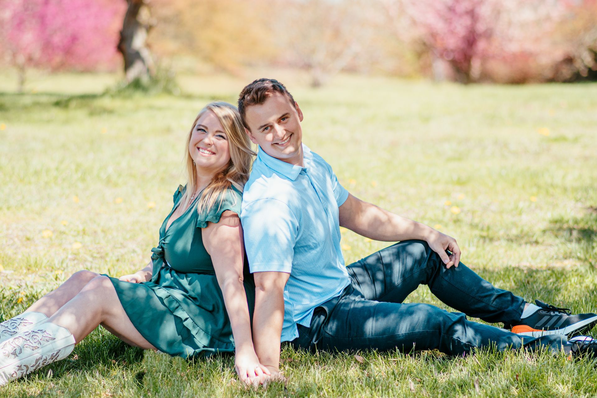 Two people sit on grass in a sunny park, leaning back-to-back with pink flowering trees behind them.