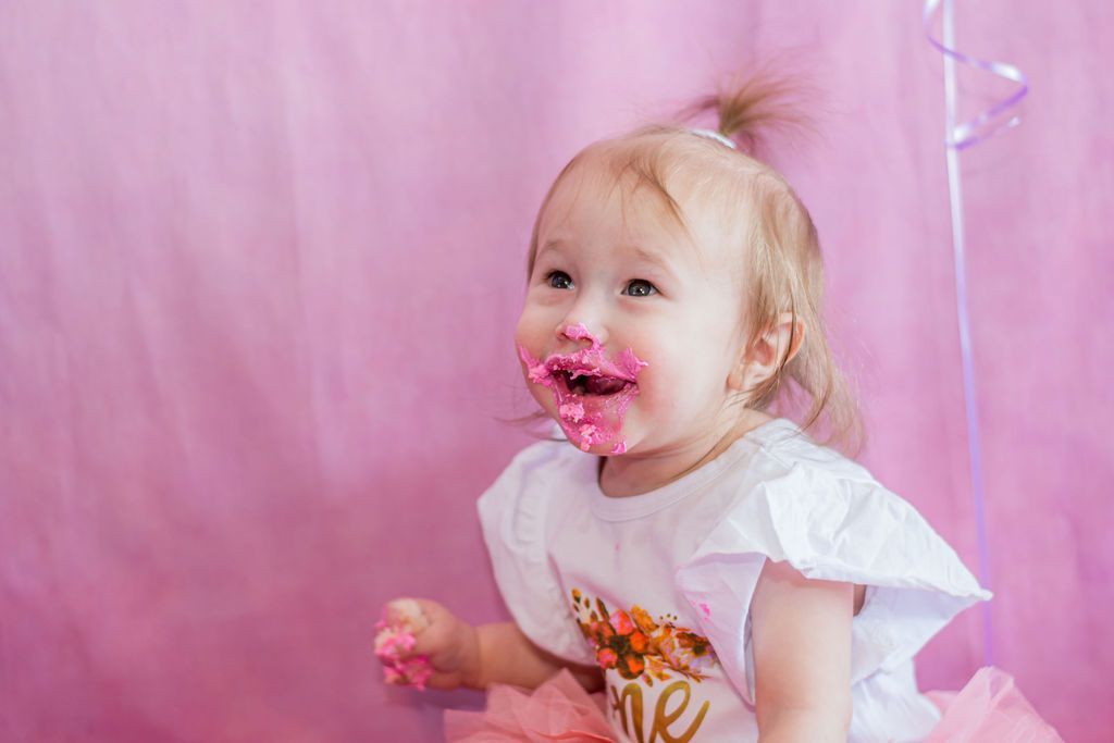 Toddler with pink frosting on face, smiling in a pink party setting with balloons.