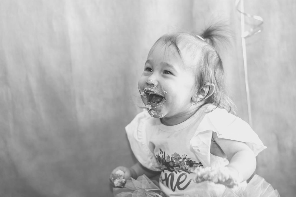 Smiling toddler with cake on face, holding a stuffed toy, in front of a plain backdrop.