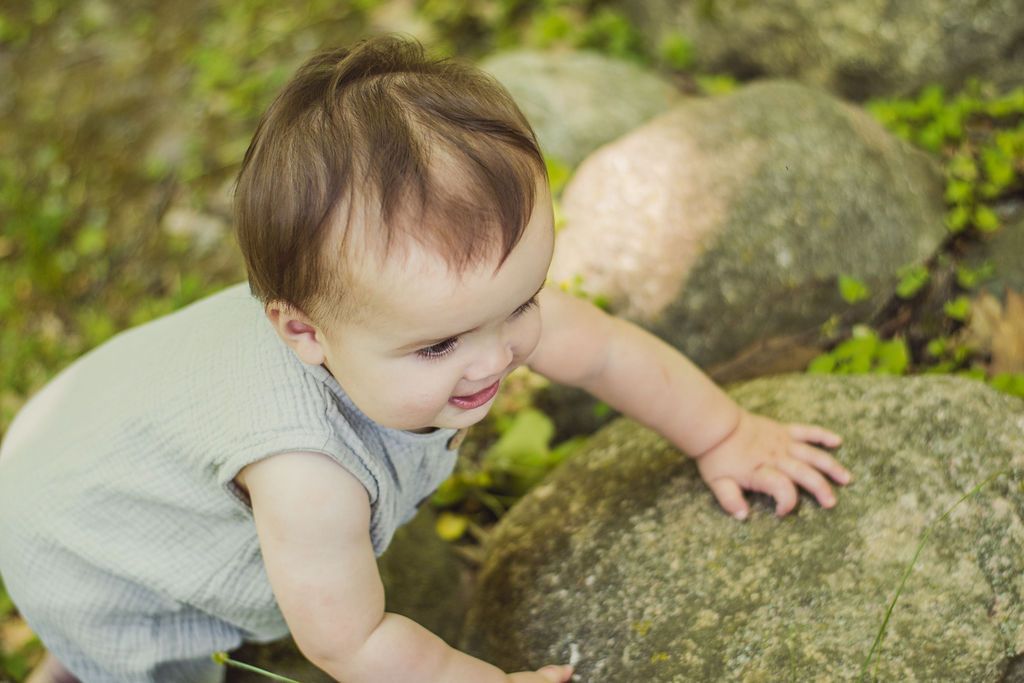 Toddler in a light gray outfit reaching toward a large rock outdoors in a grassy area