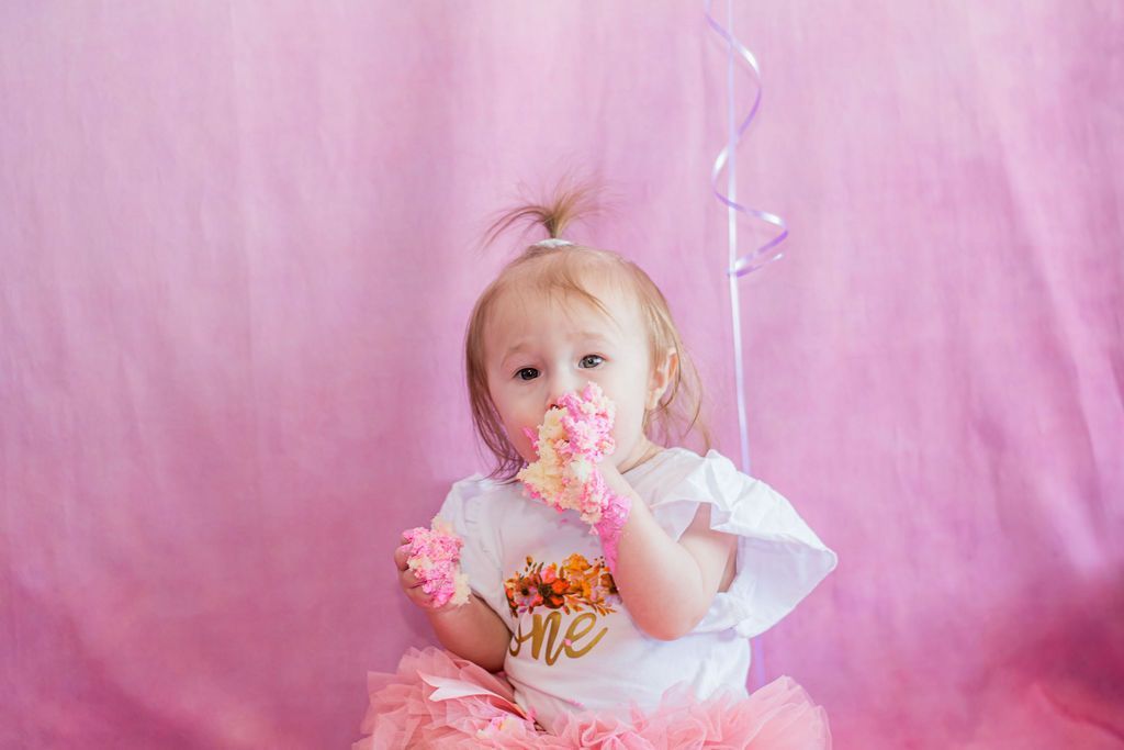 Toddler holding a pink cupcake, wearing a white shirt and pink tutu against a pink backdrop.