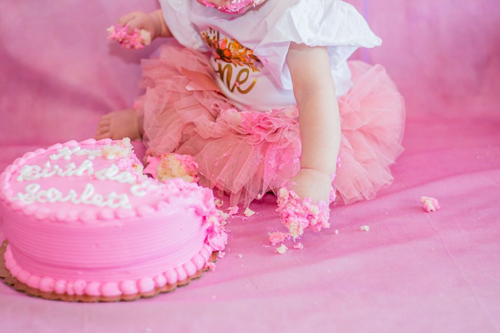 Pink birthday cake beside a child in a pink tutu on a pink floor, reaching toward the cake