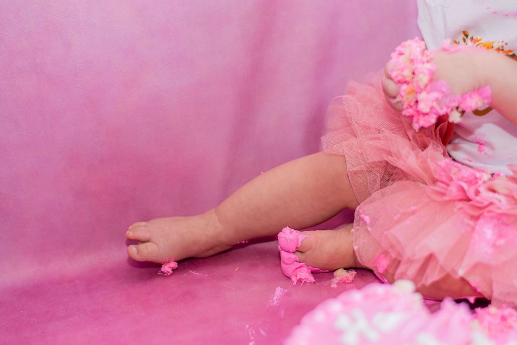 Baby legs in a pink tutu resting on a pink background, with pink flower anklets