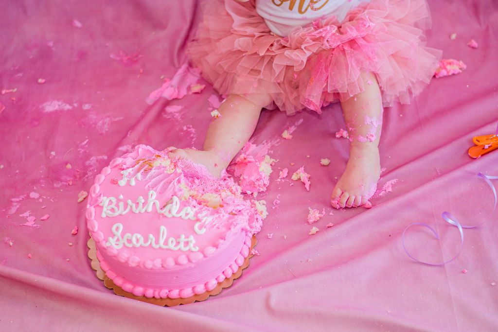 Pink birthday cake on a pink blanket beside a baby’s feet, with “Birthday Scarlett” written on top