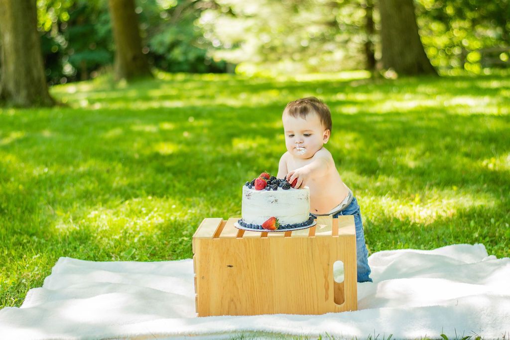 Child smiling beside a birthday cake in a wooden crate on a picnic blanket in a sunny park