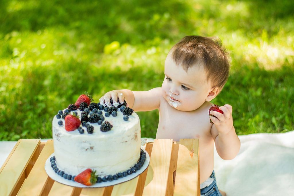 Child reaching for a berry-topped cake on a wooden table outdoors in bright sunlight