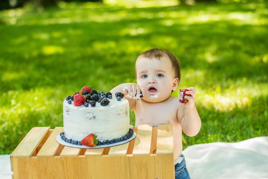 Toddler beside a berry-topped cake outdoors on a wooden crate in a sunny grassy yard