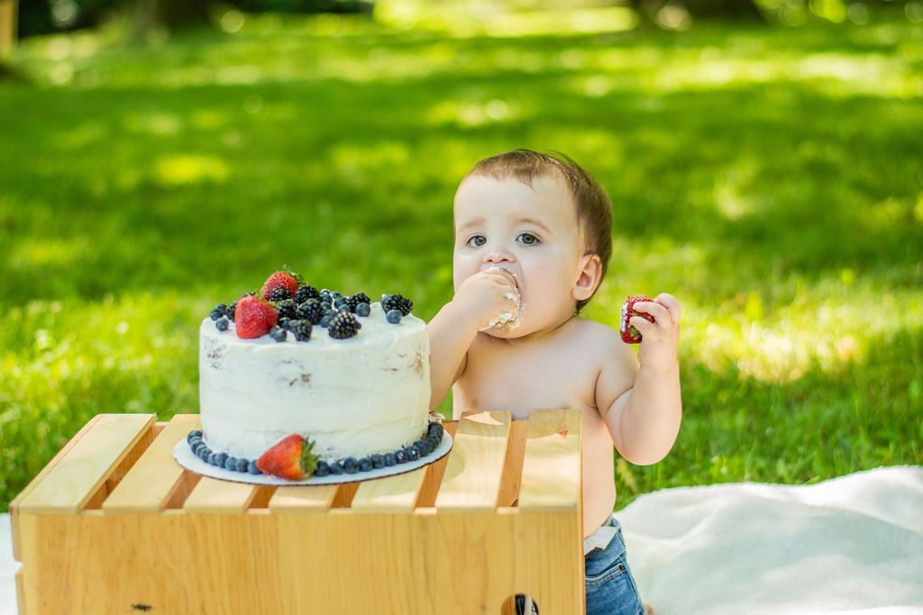 Baby reaching for a cake on a wooden crate outdoors, with green grass in the background.