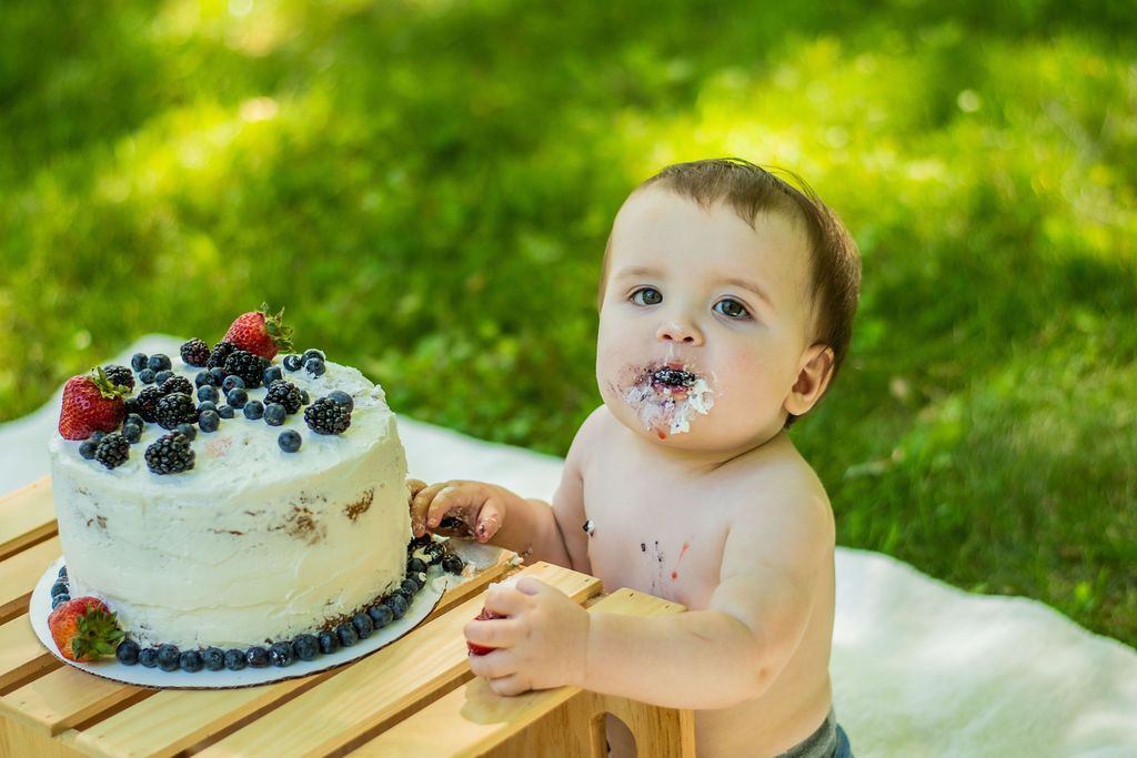 Toddler beside a berry-topped cake at an outdoor picnic table, with frosting on their face.