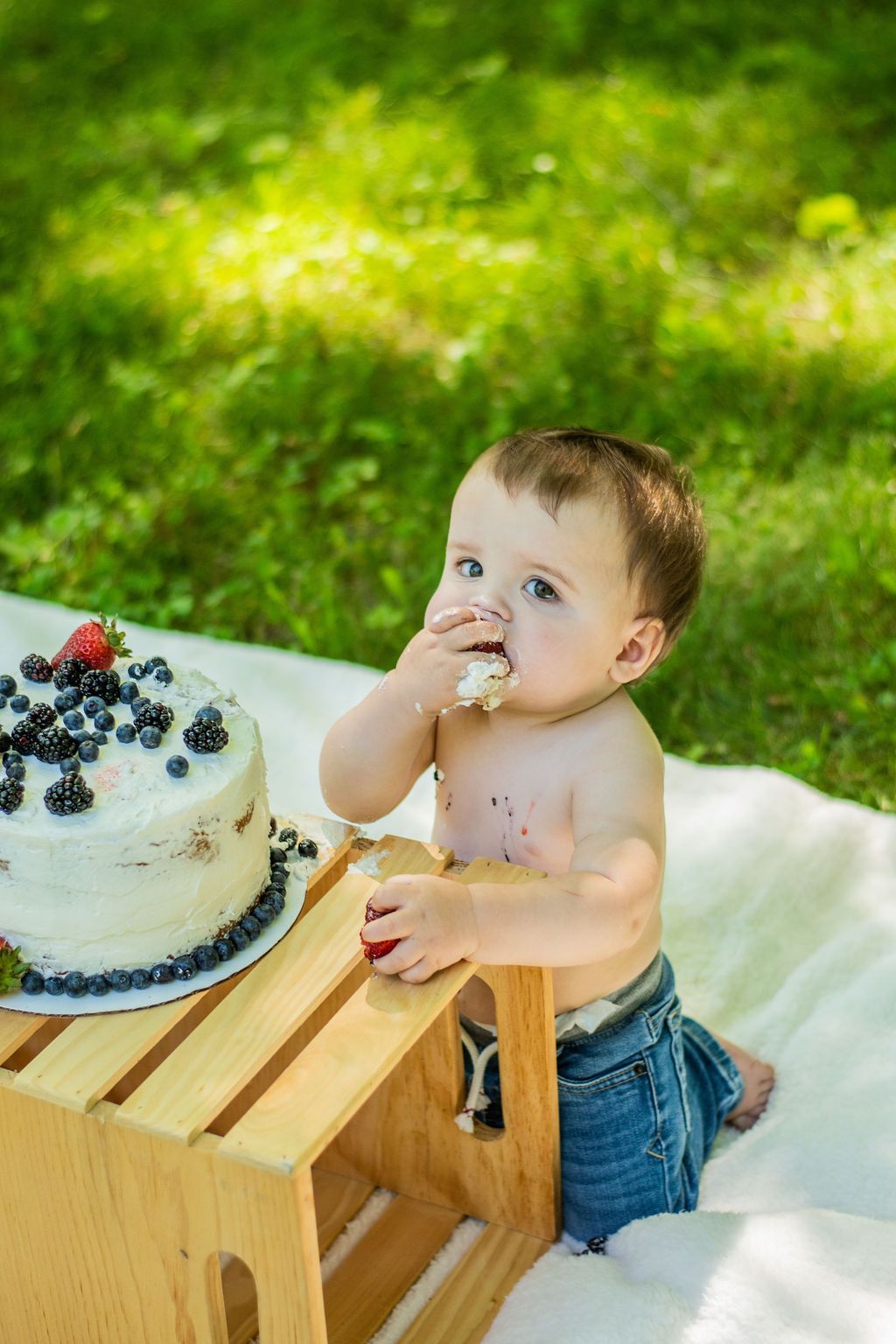 Baby beside a berry-topped cake at a picnic table on a white blanket outdoors