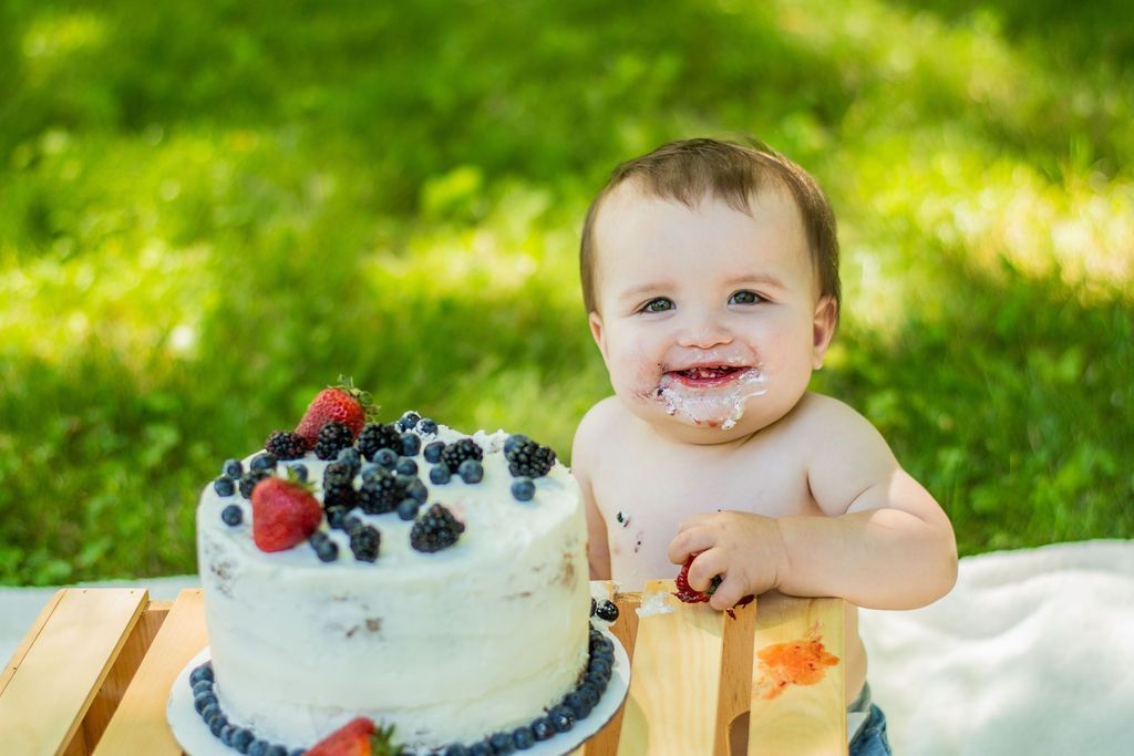 Smiling baby beside a berry-topped cake outdoors on a picnic blanket