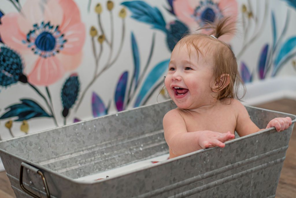 Smiling baby in a gray metal tub, with floral wallpaper in the background