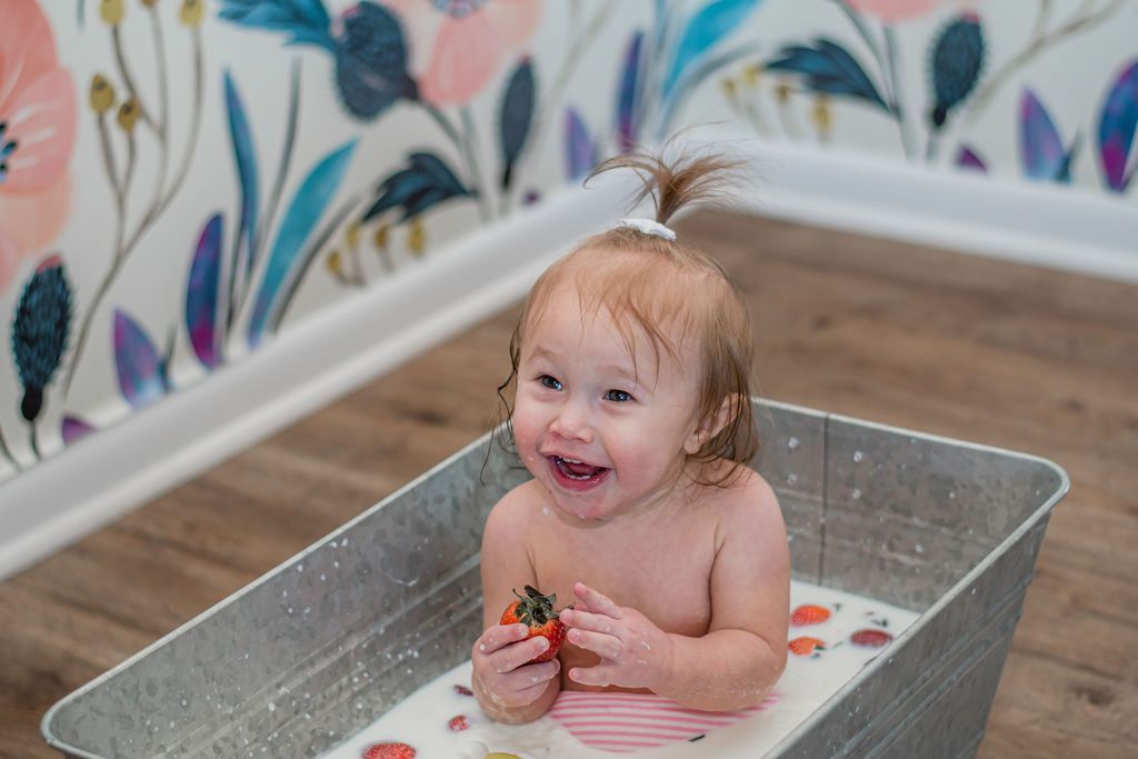 Smiling child sitting in a metal tub, holding berries, with milk and fruit on the floor and colorful wallpaper behind.