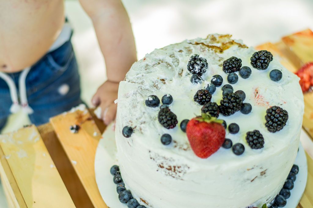 White frosted cake topped with berries on a wooden table, with a child standing nearby