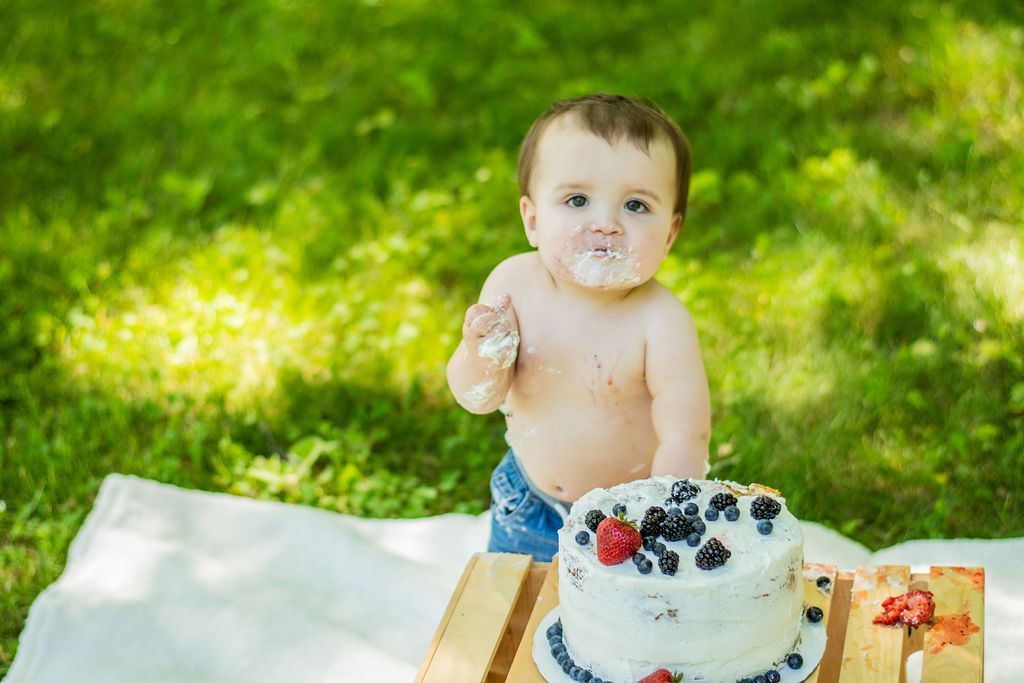 Baby at a birthday cake outdoors on grass, reaching toward the frosting with a spoon