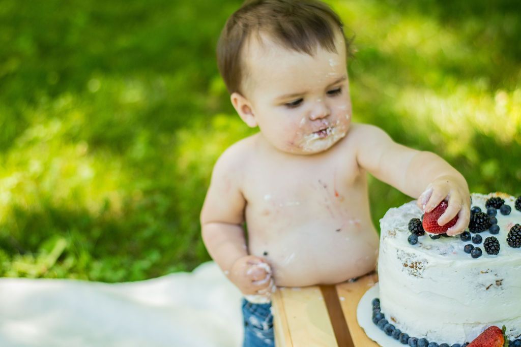 Baby touching berries on a white cake outdoors on a blanket, with frosting on their face