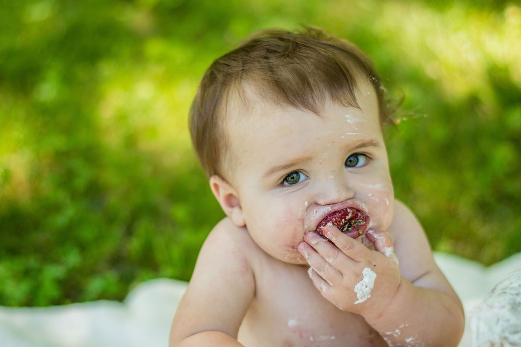 Baby on a blanket eating a cupcake outdoors on a grassy lawn