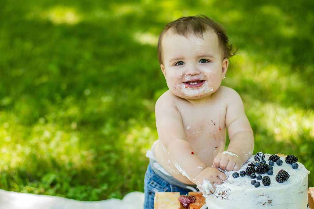 Smiling baby in jeans touching a white cake with blueberries outdoors on green grass