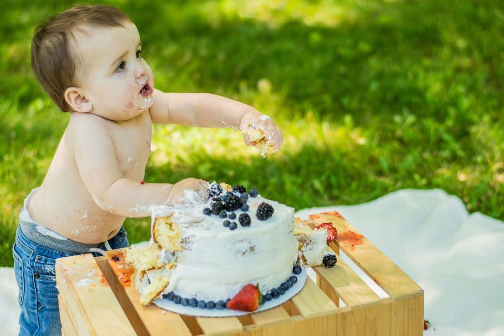 Toddler reaching into a frosted cake at an outdoor picnic table on grass