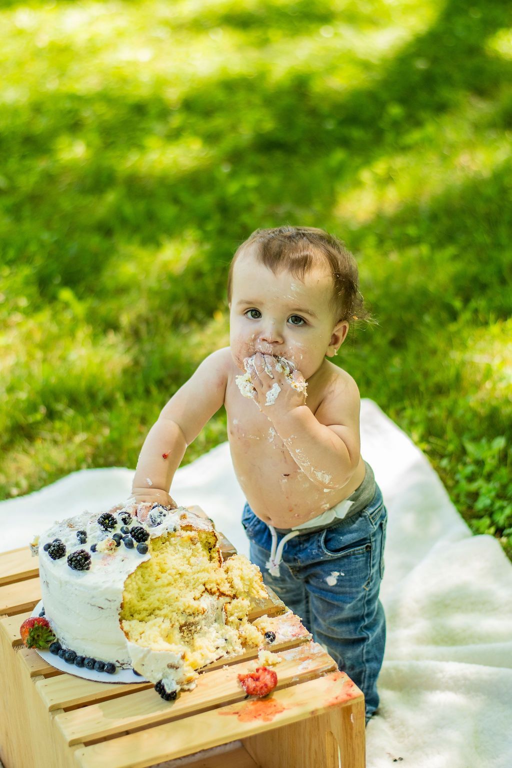 Toddler standing beside a picnic table with a cake, covered in frosting on a sunny grassy lawn
