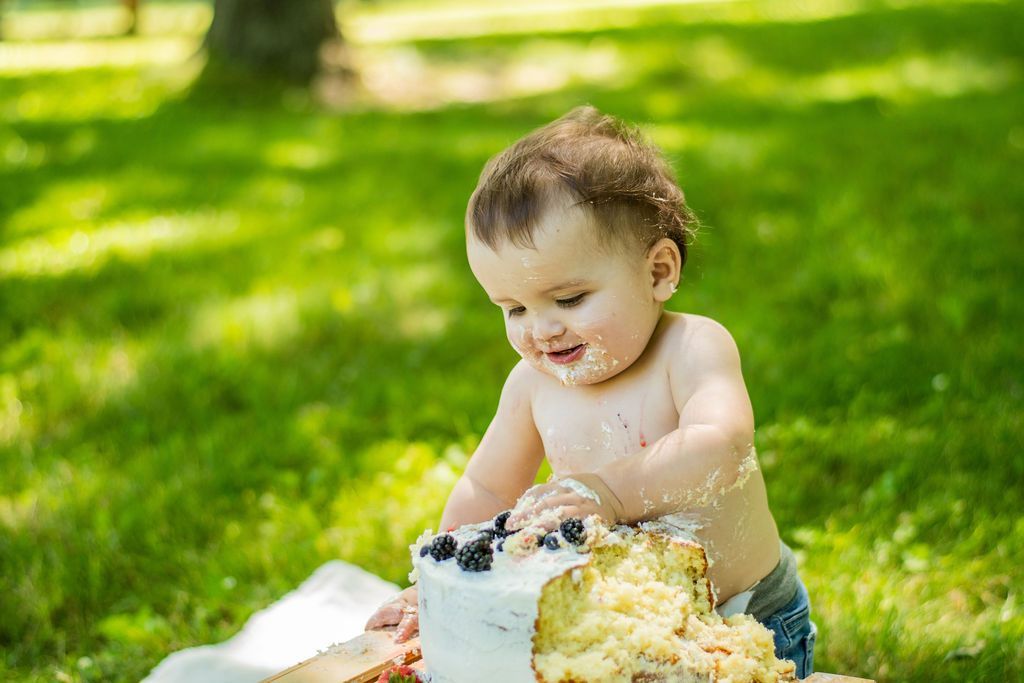 Toddler smashing a cake outdoors on green grass, smiling with frosting-covered hands