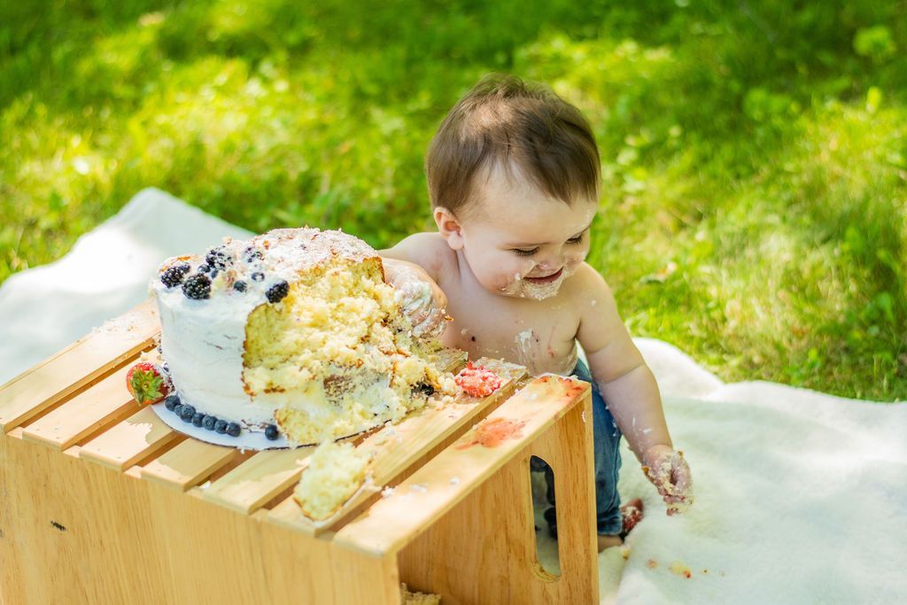 Baby beside a cake on a wooden bench outdoors, smiling on a white blanket in the grass.