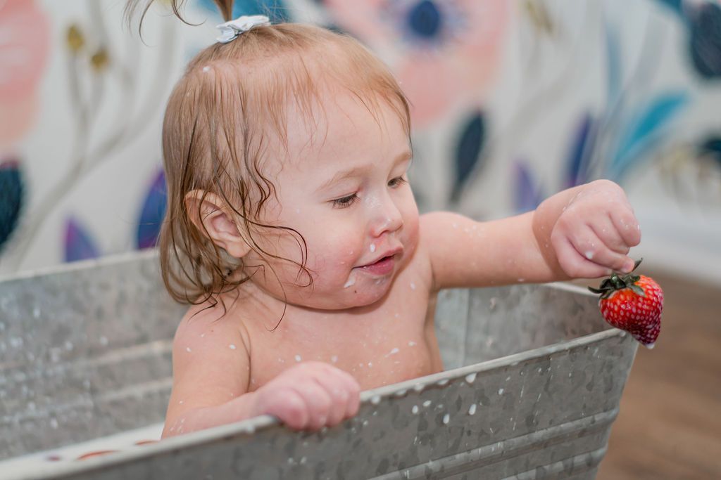 Toddler in a gray tub reaching for a strawberry, with wet hair and a focused expression