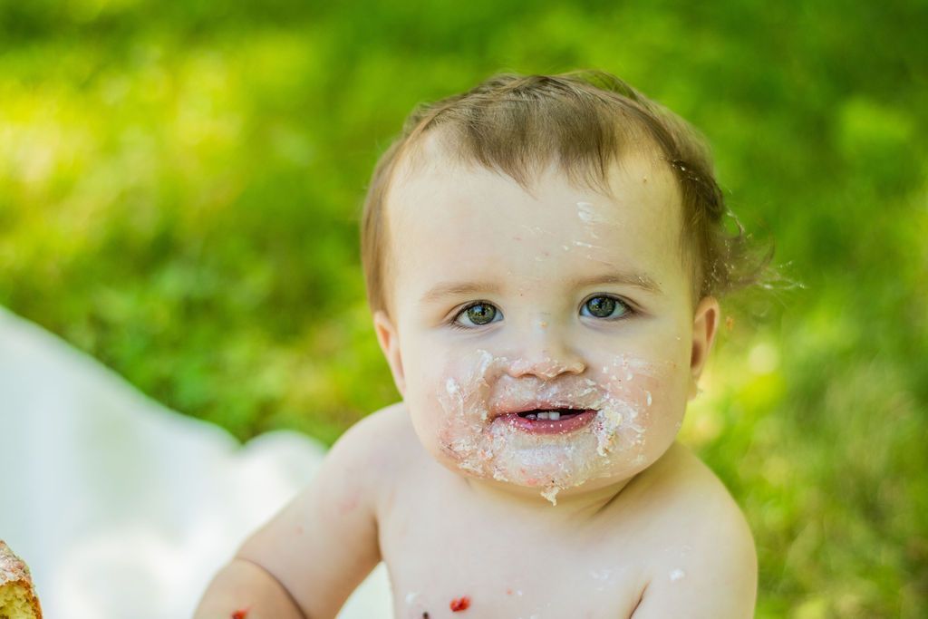 Smiling toddler with messy face sitting on a white blanket outdoors in green grass