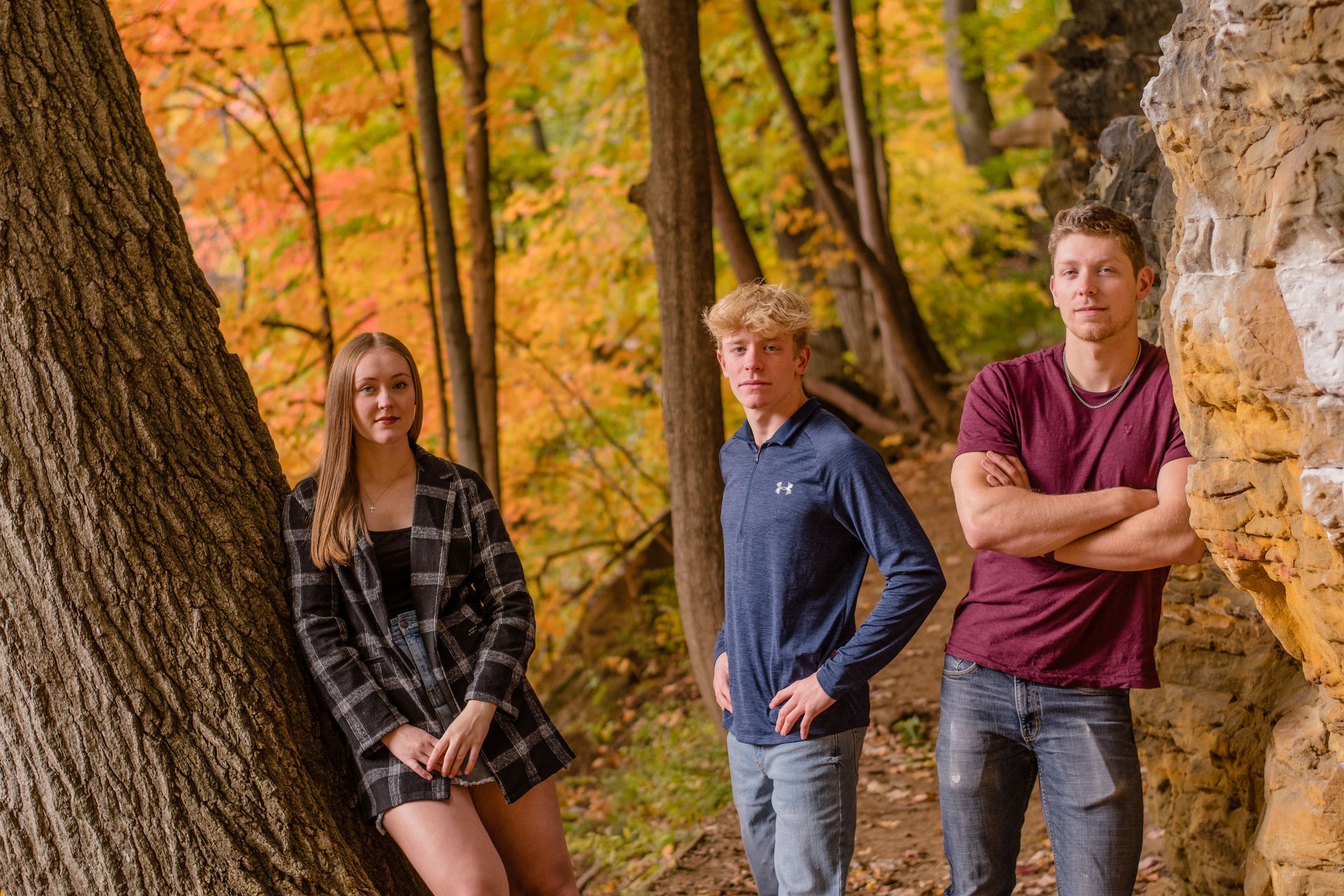 Three people posing outdoors among autumn trees and a rocky cliff.