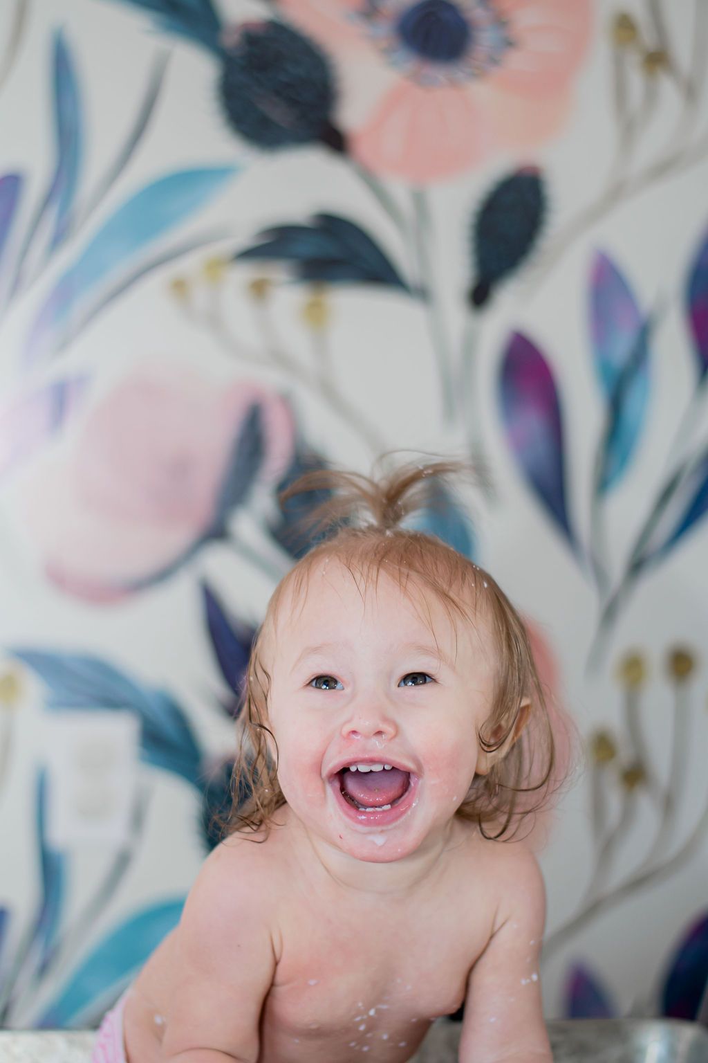 Smiling toddler with damp hair in a bath, against a pastel floral wallpaper background