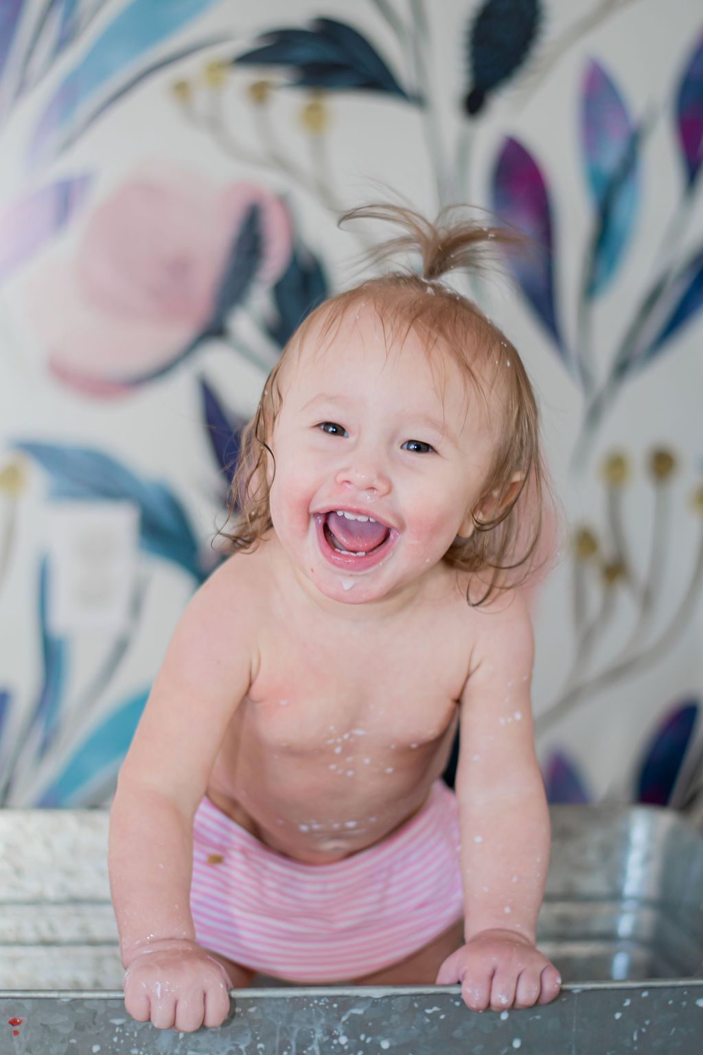 Smiling baby crawling on a gray floor, wearing pink pants, with a blurred floral mural behind.