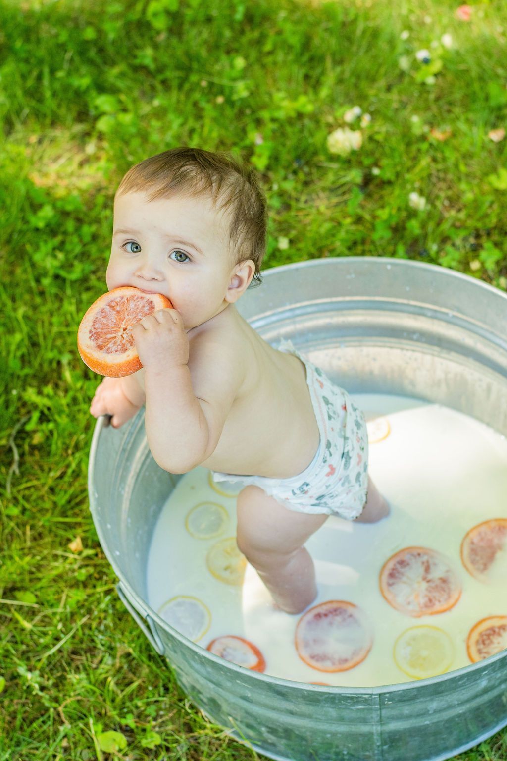 Baby standing in a metal tub of water on grass, holding a slice of orange and looking at the camera