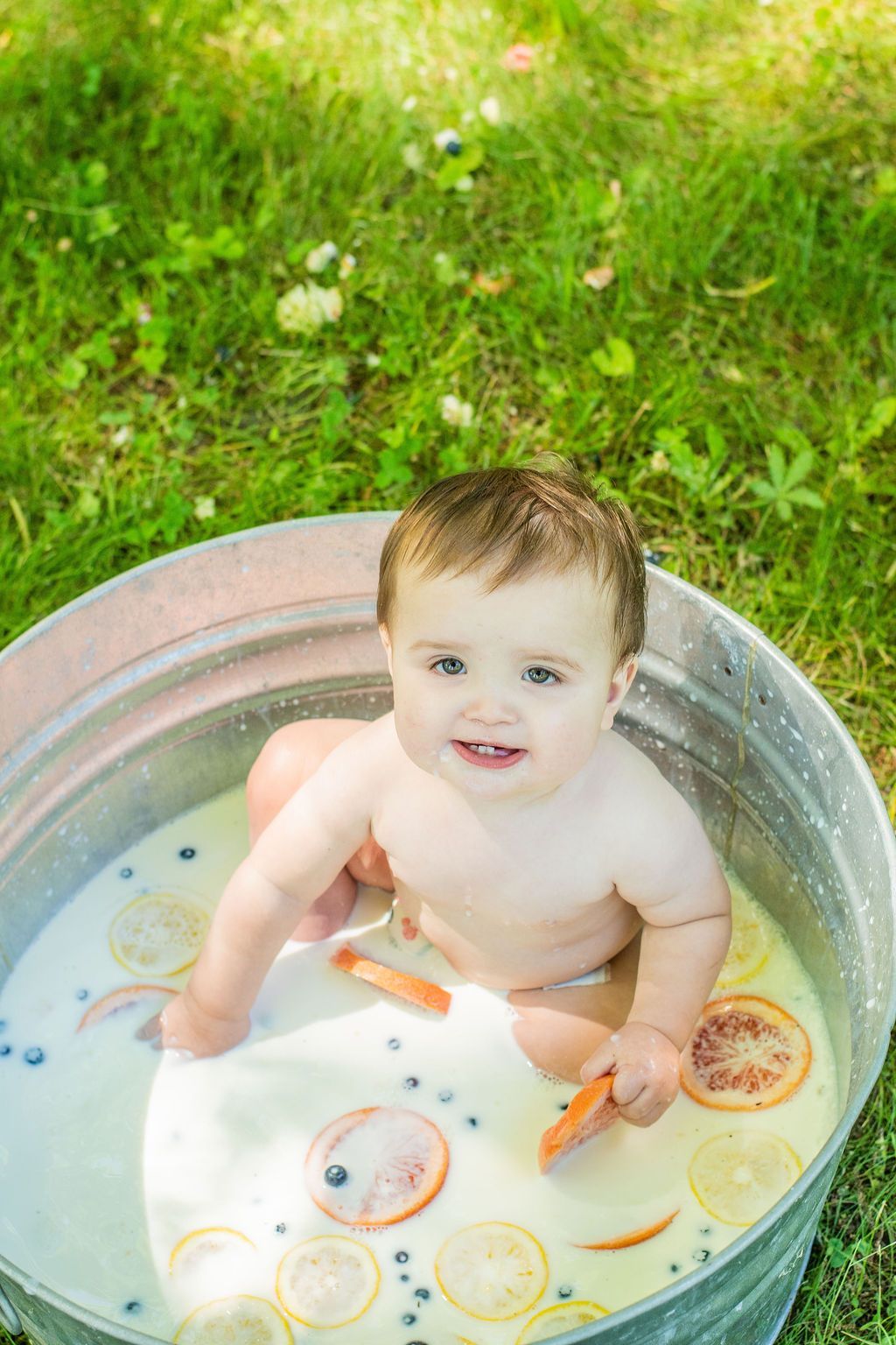 Child sitting in a metal tub of water outdoors on grass, smiling and playing with floating toys