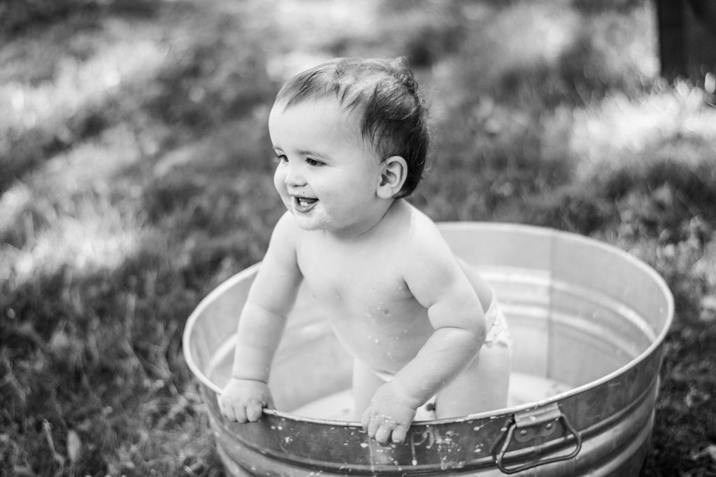 Baby sitting in a metal tub outdoors, smiling in black and white.