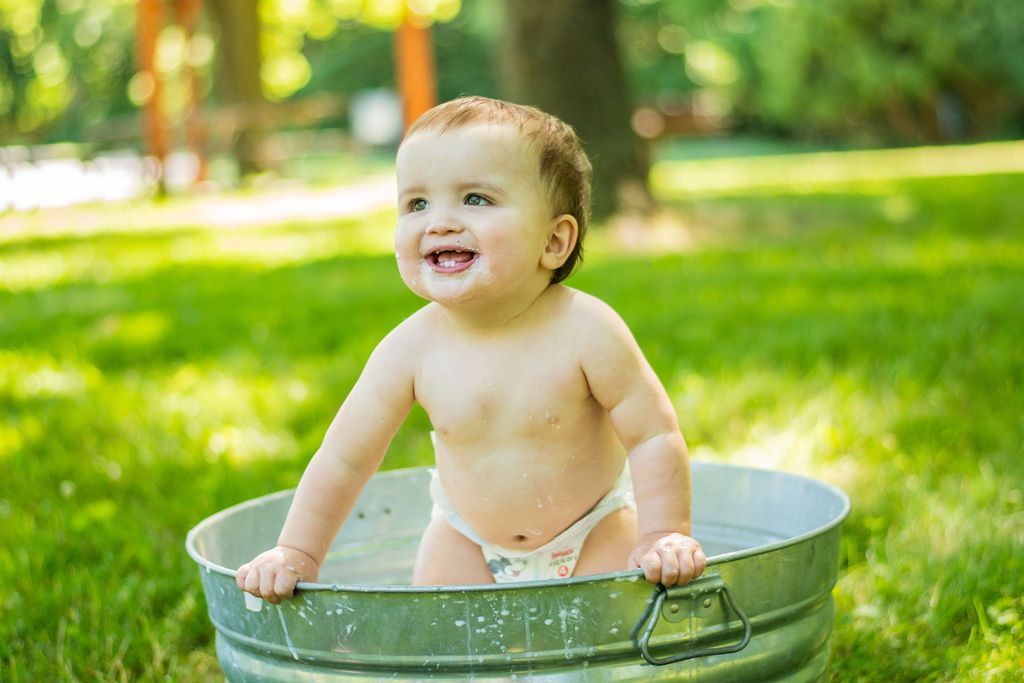 Smiling baby standing in a metal tub on green grass outdoors in sunlight