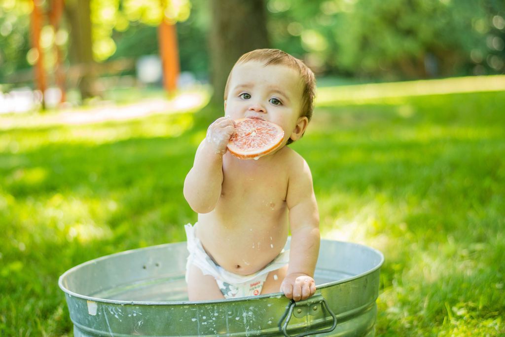 Baby sitting in a metal tub on green grass, eating a pink frosted donut outdoors.