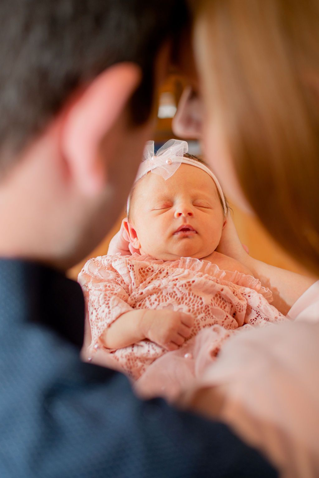 Sleeping baby in pink lace outfit, held by two adults, with a blurred warm indoor background