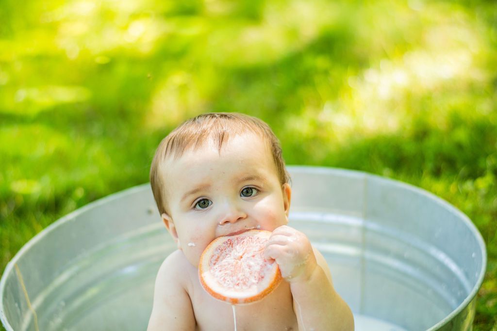 Baby sitting in a metal tub outdoors, holding and chewing a slice of citrus fruit
