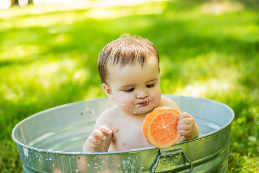 Baby in a metal tub outdoors holding an orange slice